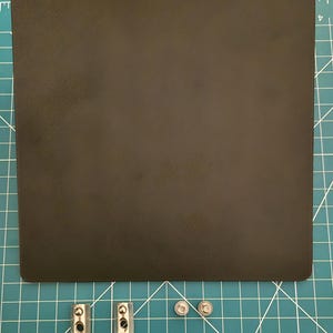 May include: A black rectangular object with rounded corners, possibly a mousepad, sits on a green grid surface. Two silver metal clamps and two small silver screws are below the object. The grid has centimetre markings.
