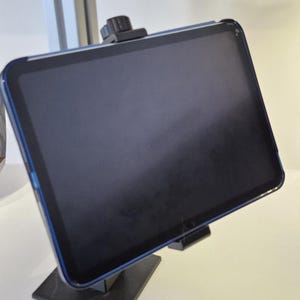 Könnte beinhalten: Ein schwarzes Tablet mit blauem Rand ist auf einem schwarzen Ständer montiert. Der Bildschirm des Tablets ist dunkel und der Ständer hat eine quadratische Basis. Das Tablet wird oben von einer schwarzen Klammer gehalten.