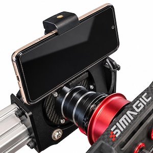 May include: A black smartphone mounted on a Simagic racing wheel base. The device is held by a black clamp attached to a carbon fiber textured plate. The assembly features a cylindrical black metal connector and a red circular component.