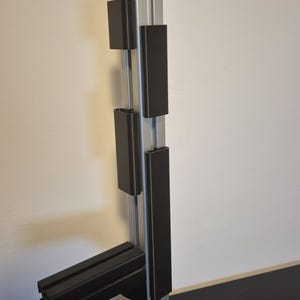 以下が含まれることがあります： 正方形のベースと垂直支持構造を備えた、黒とシルバーの金属製ディスプレイスタンド。長方形の黒いブロックが垂直支持体に取り付けられています。スタンドは、ベースから伸びる水平アームを備えています。