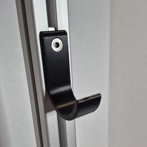 May include: A black metal hook attached to a silver metal frame. The hook is curved and has a single screw. The background is a plain white wall. The hook is designed for hanging items.