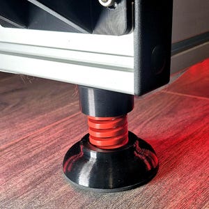 May include: A black and red adjustable foot for a piece of furniture or equipment. The foot has a black base, a red spring, and a black top section, providing stability and height adjustment. The foot is designed to be mounted on a flat surface.