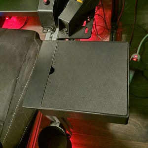 May include: A black gaming chair with a side table and cup holder. The side table is square and has a small cutout. The cup holder is round and black. The chair has a black seat and backrest.
