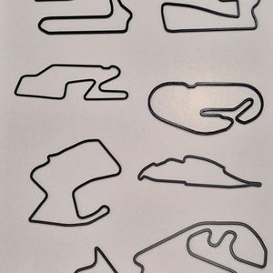 May include: Eight black outline illustrations of race tracks, each with a unique design. The tracks vary in shape and size, with sharp turns, long straights, and complex curves. The tracks are displayed on a white surface.