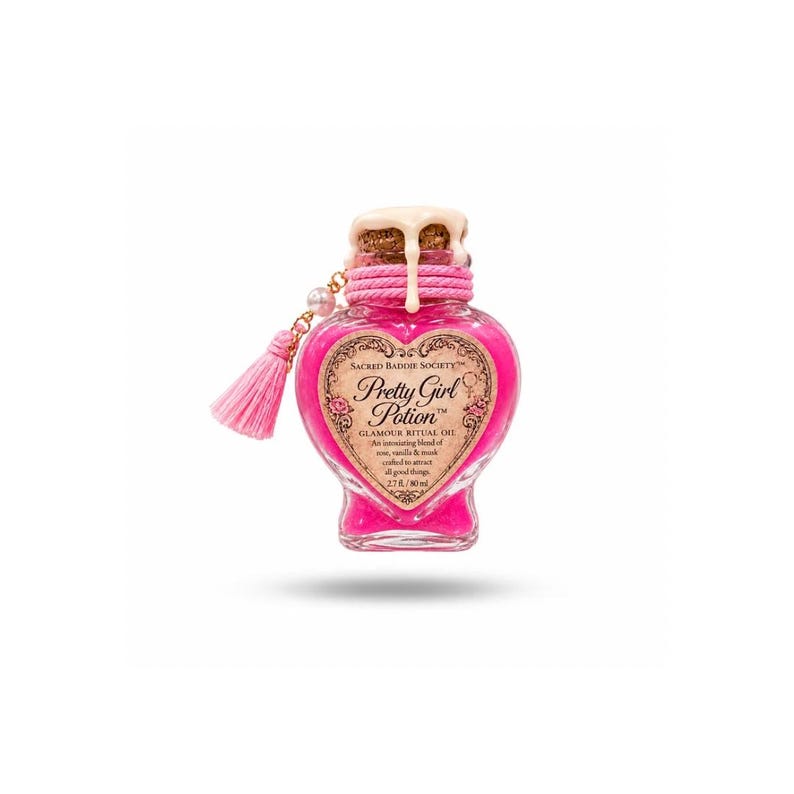 Op de afbeelding: Een hartvormige glazen fles gevuld met felroze vloeistof, met het etiket Pretty Girl Potion Glamour Ritual Oil. De fles heeft een kurk met druipende witte was, een roze koord om de hals en een roze kwastje als decoratieve hanger.