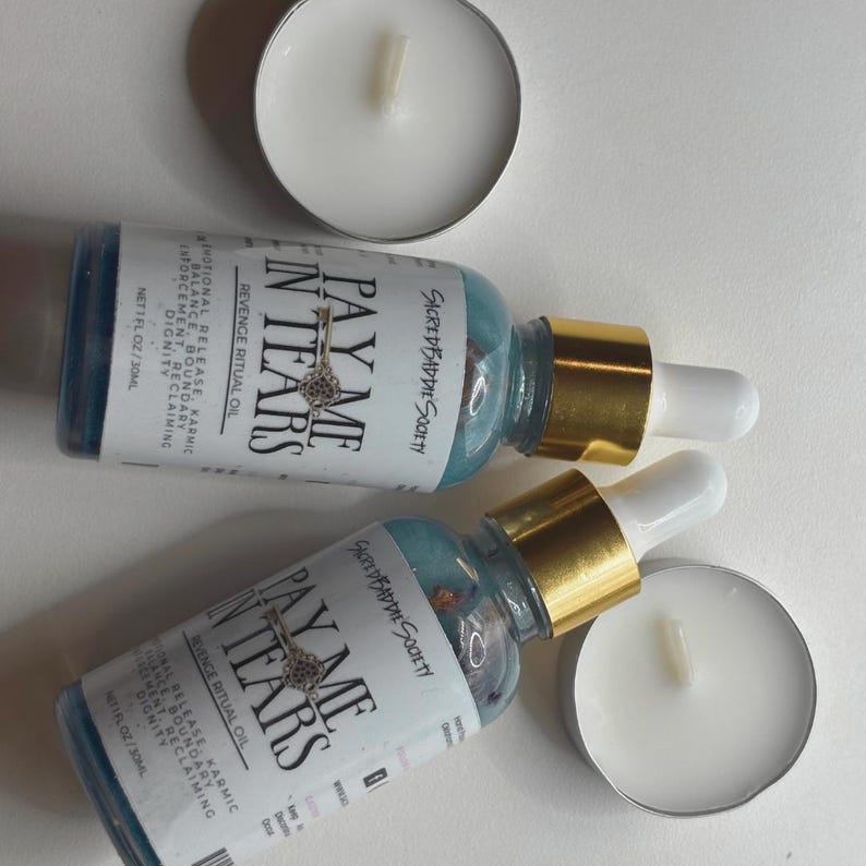 May include: Two blue glass bottles with gold-colored tops and white droppers, labeled "PAY ME IN TEARS" with a key graphic. The bottles are next to two white tea light candles. The bottles contain a blue liquid and small decorative elements.