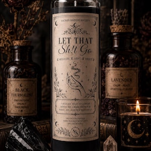 May include: A tall black pillar candle in a clear glass jar with a beige label featuring the text "LET THAT Sh!t Go", "SACRED BADDIE SOCIETY", and "I RELEASE WHAT NO LONGER SERVES ME. I MAKE ROOM FOR PEACE, JOY, AND NEW BLESSINGS." The label includes a hand illustration.