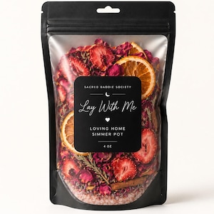 May include: A black pouch with a clear window containing a simmer pot blend. The blend features dried strawberries, orange slices, and rose petals. The label says "Lay With Me" and "Loving Home Simmer Pot".