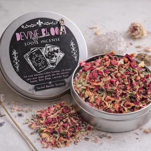 May include: A silver tin of "DEVINE BLOOM" loose incense with a key charm, next to an open tin filled with dried rose petals and herbs. The label features tarot card illustrations and the text "Sacred Baddie Society".