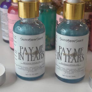 May include: Two glass bottles with gold caps and blue liquid. The labels read "Sacred Baddie Society" and "Pay Me In Tears Revenge Ritual Oil." The bottles are 1 fl oz / 30ml. Other bottles are visible in the background.