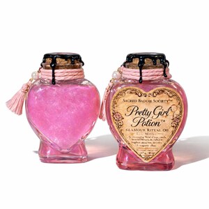 Puede incluir: Dos botellas de vidrio en forma de corazón llenas de líquido rosa. Las botellas tienen tapones de corcho con cera negra, hilo rosa y una borla. Una botella tiene una etiqueta que dice "Pretty Girl Potion" y "Glamour Ritual Oil".