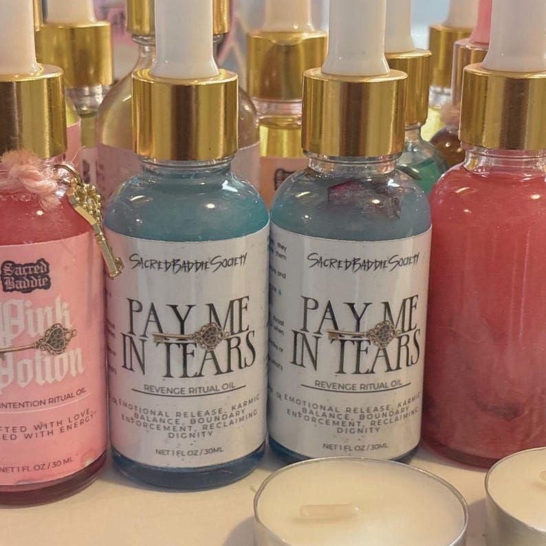 May include: Bottles of ritual oil with gold caps and white labels. The labels read "PAY ME IN TEARS" and "REVENGE RITUAL OIL". The bottles are filled with blue and pink liquids. The text "Sacred Baddie Society" is also visible.