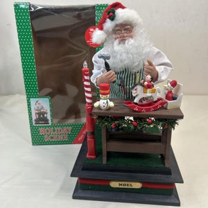 May include: A Christmas holiday scene featuring a Santa Claus figurine with a hammer, working on toys. The scene includes a rocking horse, snowman, and a sleigh. The base has the word "NOEL" and the box says "Holiday Scene".