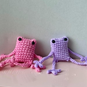 Puede incluir: Dos peluches de rana tejidos a ganchillo a mano, uno en rosa brillante y otro en morado claro. Cada rana presenta un cuerpo rectangular, extremidades largas y delgadas, y dos pequeños ojos de cuentas de plástico negro. Se nota la textura.