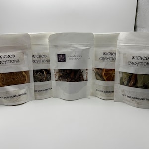 Può includere: Cinque sacchetti bianchi con finestre trasparenti, ciascuno etichettato "WICKED CREATIONS" o "PUMPKIN SPICE SIMMER POT". I sacchetti contengono erbe e spezie essiccate, tra cui fette d'arancia e foglie di alloro. Il testo "NOT FOR CONSUMPTION" è stampato.