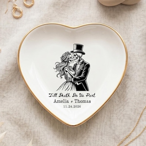 Op de afbeelding: Een wit hartvormig keramieken schaaltje met een goudkleurige rand. In het midden staat een zwarte afbeelding van twee skeletten die elkaar omhelzen, met de tekst "Till Death Do Us Part", de namen "Amelia + Thomas" en de datum "24-11-2026".