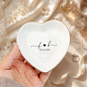 May include: White heart-shaped ceramic trinket dish with a gold rim. The dish features the initials "f & b" with a heart symbol and the date "08.16.2025" in black script. A ring and earrings are in the background.