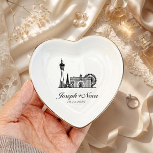 May include: Heart-shaped white ceramic trinket dish with a gold rim. The dish features a black graphic of Las Vegas landmarks and the names "Joseph + Nova" with the date "10.14.2025".