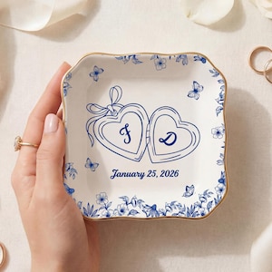 Może przedstawiać: Biała ceramiczna patera na pierścionki ze złotą obwódką, zdobiona niebieskimi wzorami kwiatowymi i motylami. Dwa medaliony w kształcie serca z inicjałami "F" i "D" są wyśrodkowane, z datą "25 stycznia 2026" poniżej.