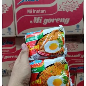 May include: Two packages of Mi Goreng instant noodles are held in front of a stack of boxes. The packages show a photo of noodles with an egg and vegetables. The boxes are red and white.