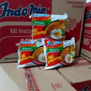 May include: Several packages of Indomie Mi Goreng instant noodles are stacked on cardboard boxes. The packaging is red, green, and white, with images of noodles and eggs. The boxes are also red with the Indomie logo.