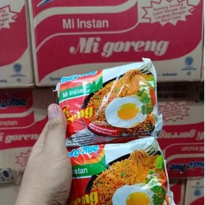 May include: Two packages of Mi Goreng instant noodles are held in front of a stack of boxes. The packages feature a photo of noodles with a fried egg. The packaging is red, white, and green with the words "Mi Instan" and "Mi Goreng".