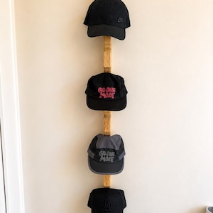 May include: A vertical wooden wall-mounted rack holding four baseball caps. The top cap is black with a small logo, the second is black with pink text reading GO ONE MORE, the third is grey and black with grey text reading GO ONE MORE, and the bottom is black with an LA logo.