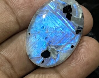May include: Oval-shaped moonstone gemstone with a blue and white iridescent surface. The stone has black inclusions and is held in a hand. The gemstone is polished and ready for jewelry making.