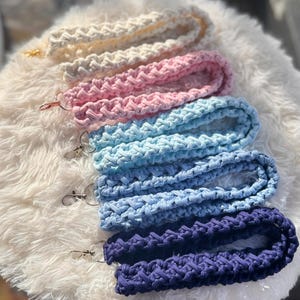 Puede incluir: Una colección de cordones hechos a mano en varios colores, incluyendo crema, rosa, azul claro, azul medio y azul marino. Cada cordón presenta un diseño tejido y un cierre metálico para sujetar llaves o una tarjeta de identificación. Los cordones se exhiben sobre una superficie blanca y esponjosa.