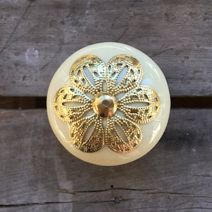 Peut inclure: Un bouton en céramique blanche avec un motif de fleur en métal doré. La fleur a un motif délicat et complexe.