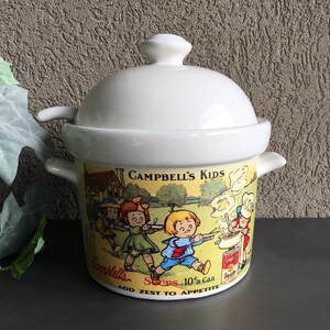 May include: White ceramic soup tureen with a lid and two spoons. The tureen features a colorful illustration of children playing with a can of Campbell's Soup. The text on the tureen reads "Campbell's Kids Add Zest to Appetite Soups 10¢ a Can".