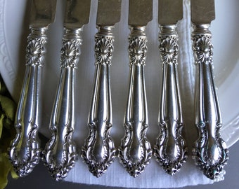 May include: Set of six silver butter knives with ornate handles. The knives are arranged in a row on a white surface.
