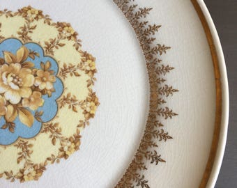 May include: White ceramic plate with a gold rim and intricate floral designs. The plate features a central floral medallion in yellow, blue, and brown, surrounded by a decorative border. The plate has a diameter of approximately 10 inches.