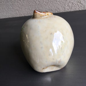 May include: A ceramic apple sculpture with a white glaze and brown accents. The apple is slightly misshapen and has a glossy finish.