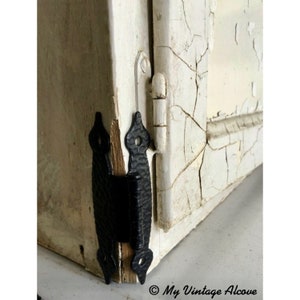 Puede incluir: Primer plano de una puerta de armario de madera pintada de blanco con bisagras de metal negro. La puerta tiene un acabado desgastado con grietas en la pintura. Las bisagras son decorativas y tienen una superficie texturizada.