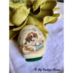 May include: A white ceramic egg with a vintage illustration of a dog and a rabbit. The egg is sitting on a green velvet base.