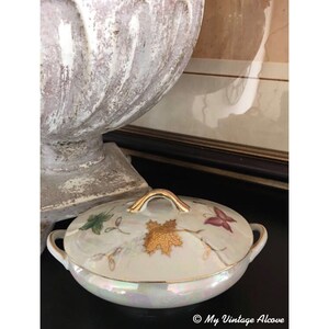 May include: A small, oval-shaped porcelain dish with a lid. The dish is decorated with a gold leaf design and a butterfly. The lid has a gold handle.