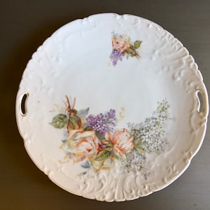 May include: White porcelain serving platter with a raised rim and two handles. The platter is decorated with a floral design featuring pink and white roses and purple lilac.
