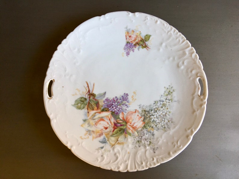 May include: White porcelain serving platter with a raised rim and two handles. The platter is decorated with a floral design featuring pink and white roses and purple lilac.