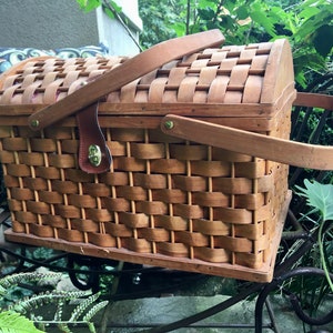 Large Vintage Picnic Basket and Dishes Set for 4. Chains support Lid. Red Fabric Interior. 4 White Enamel Cups and Plates. 12 inches high not including handles. 17.5 inches wide. Pre-owned with a couple breaks in wood. Some chipping of enamel dishes.