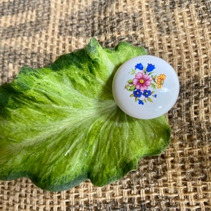May include: A white ceramic knob with a floral design featuring pink, blue, and yellow flowers. The knob is on a green leaf.