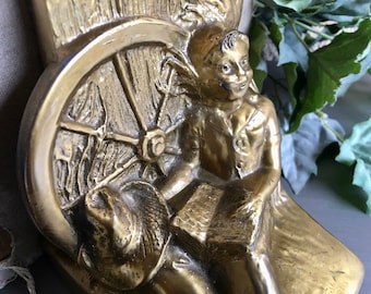 May include: A pair of gold-tone bookends featuring a sculpted figure of a child sitting on a wheel. The child is holding a book and has a smiling expression.