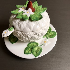 Puede incluir: Azucarero y tapa de cerámica blanca con forma de calabaza con hojas verdes y flores blancas. El tazón se asienta sobre un plato a juego con hojas verdes y flores blancas.