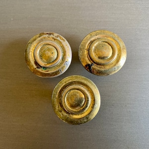 Könnte beinhalten: Drei goldfarbene runde Schrankknöpfe mit konzentrischem Kreisdesign. Die Knöpfe haben einen leicht abgenutzten, antiken Look.