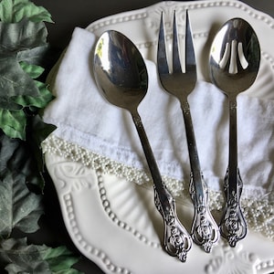 以下が含まれることがあります： スプーン、フォーク、穴あきスプーンの3つのヴィンテージシルバーサービング用具のセット。装飾的な縁と白いリネンナプキンが付いた白いプレートの上に置かれています。ハンドルには、華やかなフローラルデザインが施されています。