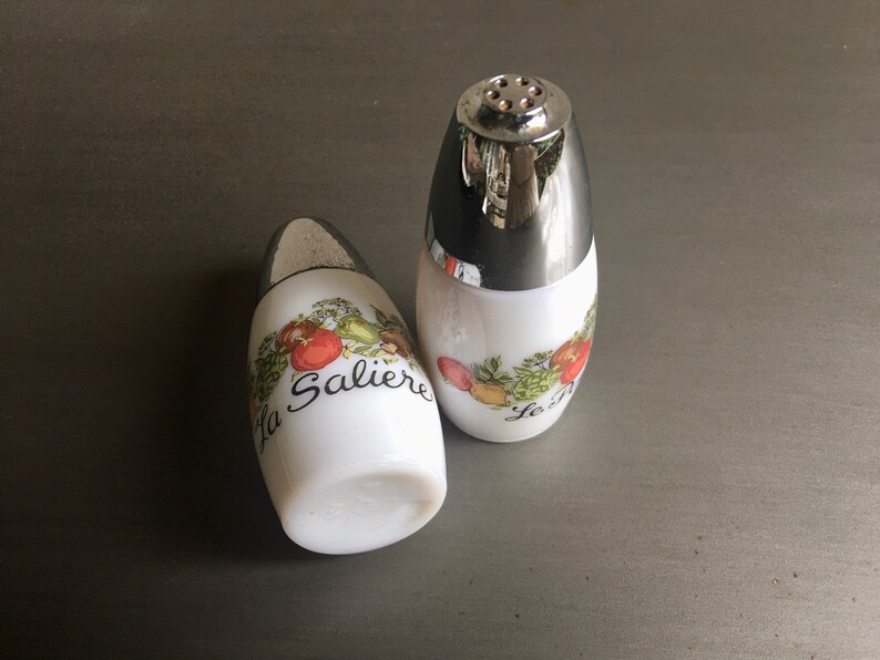 Vintage White Milk Glass Salt and Pepper Shakers crafted by Gemco Ware. Adorned with elegant French inscriptions, La Saliere for salt and Le Poivrier for pepper, in black cursive lettering. Backstamp dates Shakers between the years 1960 and 1980.