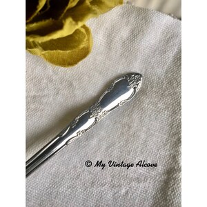 May include: Close-up of the ornate, silver handle of a vintage spoon. The handle is decorated with a floral design.