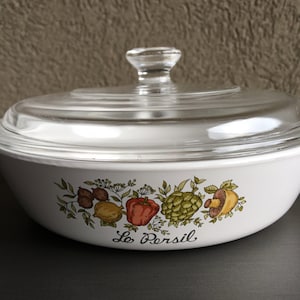 May include: White casserole dish with a clear glass lid. The dish features a colorful vegetable design with the text "Le Persil" in black script. The lid has a clear glass knob. A vintage kitchenware item.
