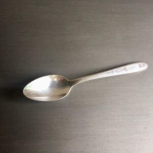 May include: A silver spoon with a decorative pattern on the handle. The spoon is lying on a dark gray surface.