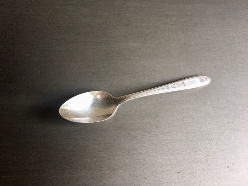 May include: A silver spoon with a decorative pattern on the handle. The spoon is lying on a dark gray surface.